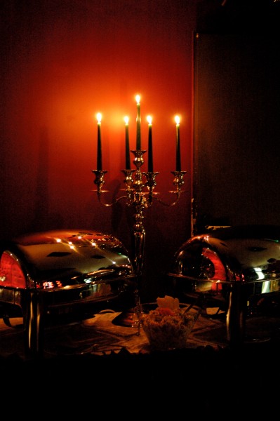 Dwa błyszczące, metalowe bemary z podgrzewaczami, ustawione na stole z dekoracją, oświetlone świecami w srebrnym, pięcioramiennym świeczniku na tle czerwonej ściany. Oświetlenie tworzy ciepłą...